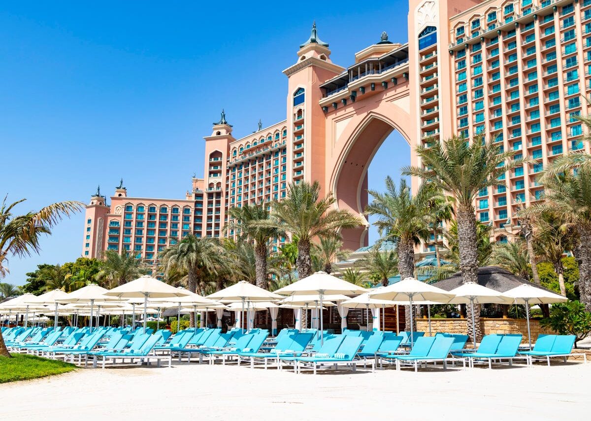 Resort de luxo em Dubai com arquitetura imponente, palmeiras, praia de areia branca e espreguiçadeiras à beira-mar.