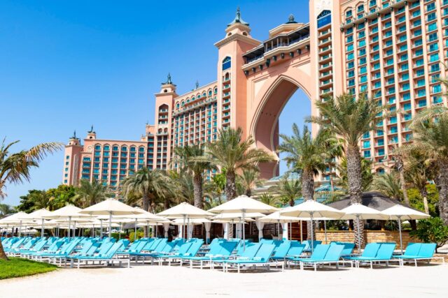 Resort de luxo em Dubai com arquitetura imponente, palmeiras, praia de areia branca e espreguiçadeiras à beira-mar.